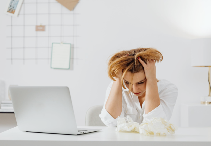 An image of a woman sitting in front of a laptop holding her head